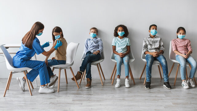 In a clinic, a medical worker vaccinates a preteen girl as a diverse group of kids wait for their turn. Each child is wearing a face mask, ensuring safety during the vaccination process.