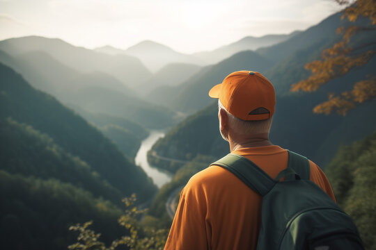 Active Senior Man Wearing an Orange Hat and Backpack Looking Out Over a Deep River Valley and Forest-Covered Mountains at Sunrise. Retirement travel, hiking, and healthy aging concept