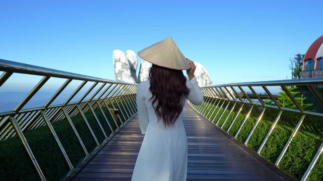 4k slow motion video of Asian woman wearing white dress, Vietnamese culture with traditional conical hat on Golden Hand Bridge, on a clear morning with blue sky, Danang city, Vietnam.