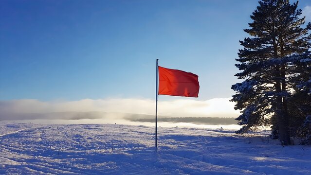 A vibrant red flag stands tall in a vast snowy winter landscape under a clear blue sky with distant fog and sunlit pine trees
