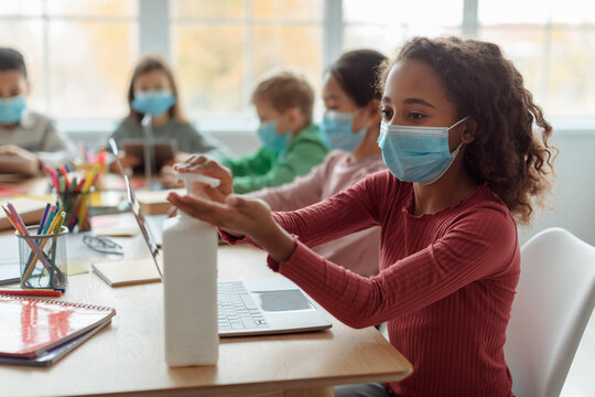 In a classroom setting, a black schoolgirl wearing a face mask uses hand sanitizer while sitting with classmates. Everyone practices hygiene during Covid-19 for safety and health. - Powered by Adobe
