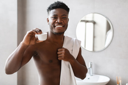 A man stands in a bathroom wrapped in a towel, smiling as he holds a jar of moisturizer cream up to the camera. His confidence shines as he promotes skin and body care.