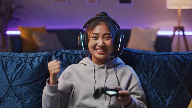 Excited Gamer Celebrating Victory - A young woman wearing a hoodie and colorful LED headphones enthusiastically celebrates a gaming victory, holding a game controller in her hands.