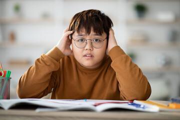 A chubby Asian teenager wearing glasses is sitting at a table covered with books and notebooks. He looks stressed, touching his head and staring at the camera, indicating a need for help.