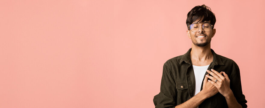 Young man stands with a warm smile, hands over his heart, dressed casually in a black jacket and a light shirt. The soft pink backdrop creates a cheerful and uplifting atmosphere.