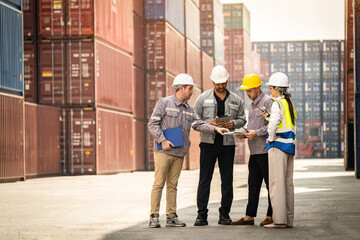 Fototapeta premium A group of workers are standing in a warehouse, looking at a tablet. They are wearing hard hats and safety vests