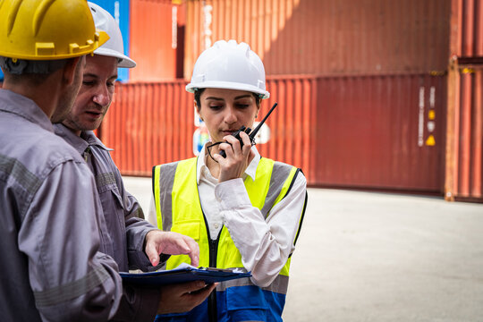 A woman in a safety vest is talking on a walkie talkie while two men look on. The woman is wearing a yellow vest and a hard hat, indicating that she is a worker in a construction or industrial setting - Powered by Adobe