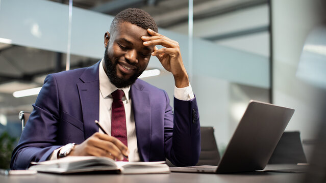 Positive wealthy young black manager working at modern office, sitting at workdesk in front of laptop, taking notes and smiling, panorama with copy space. Business opportunities concept