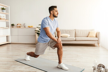 A young man is performing a lunge stretch on a yoga mat in a well-lit living room. The space features light colors, a couch, and exercise equipment, creating a calm atmosphere for fitness.