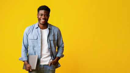A young man stands confidently, holding a laptop and smiling warmly. He is dressed casually in a blue shirt and white t-shirt against a vibrant yellow backdrop, showcasing a happy mood.