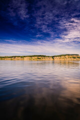 A vertical, symmetrical-style landscape of a forested coastal bluff, reflected in the smooth, calm...