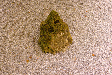 A top-down, minimalist photograph of a single, moss-covered rock placed in a bed of finely raked white and grey gravel, evoking a sense of calm and zen.