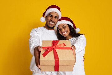 A cheerful couple wearing red Santa hats smiles as they hold out a gift wrapped with a red ribbon. The bright yellow background adds to the festive atmosphere.