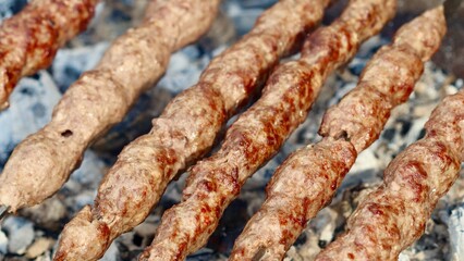 Close-up of several long, minced meat kebabs, lula or kofta, grilling on metal skewers over hot charcoal embers.