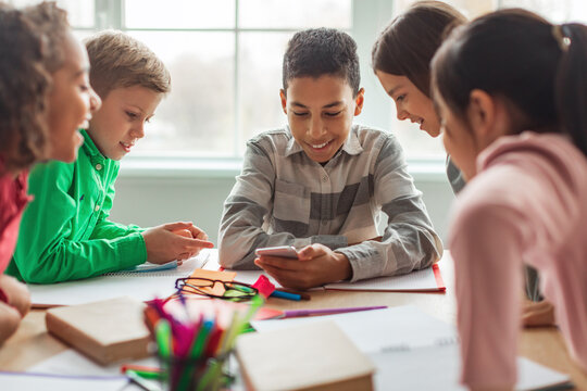 In a modern classroom, a Black schoolboy happily uses his phone while surrounded by multiracial classmates. They enjoy sharing excitement during their break, engaged in technology and education.