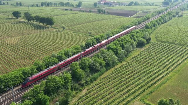 A fast-moving red high-speed train blazes across lush vineyards, captured from above. The motion contrasts with the peaceful countryside.
