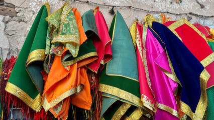 A vibrant assortment of traditional ethnic textiles, likely Indian or Middle Eastern garments such as lehenga cholis, dupattas, or religious deity clothing, hangs on display. The fabrics are a riot of