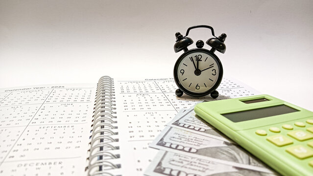 A black clock, green calculator, and dollar bills placed on an open calendar representing the concept of time management, financial planning, budgeting, and saving for future goals.
