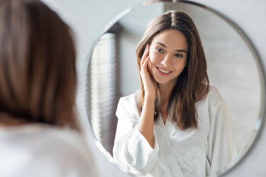 In a bright bathroom, a young woman smiles at her reflection in a round mirror. She touches her face gently, appreciating her smooth skin and the moment of self-care.