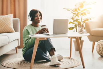 A woman sits on the floor of her living room, wearing headphones and smiling. She holds a coffee...