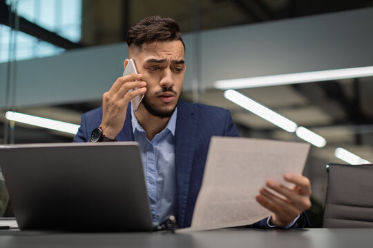 In a modern office, a young arab manager with a worried expression is reading financial documents while engaged in a phone conversation. He appears puzzled by the report he is reviewing.