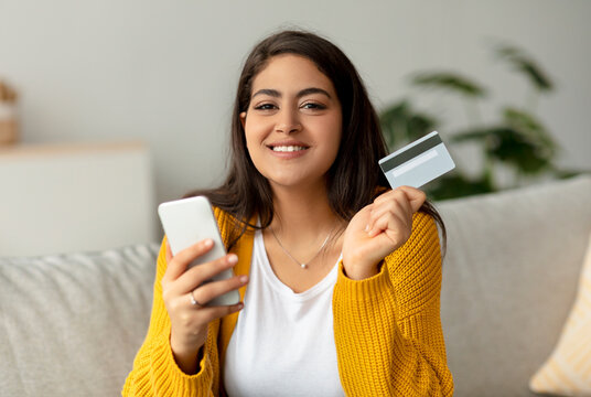 A cheerful Muslim lady is making an online payment using her smartphone and credit card. She is seated on her couch and looks happy while engaging in shopping from home. - Powered by Adobe