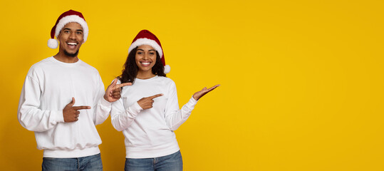 Two cheerful individuals wearing white sweaters and Santa hats stand against a bright yellow background, pointing with smiles. They joyfully celebrate the Christmas season together.