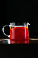 Glass teapot with hot red herbal tea on wooden table in dark background