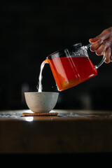 Pouring hot herbal tea from glass teapot into ceramic cup on dark background
