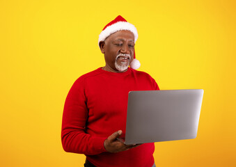 A happy senior black man wears a Santa hat and red sweater as he enjoys online shopping for...