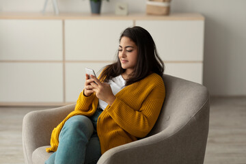 A positive Arab woman sits comfortably in an armchair, using her cellphone to check social networks. She enjoys her time at home, engaging in various activities on the app.