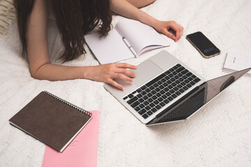 Teenage girl with laptop and notebook working online on your bed in bedroom