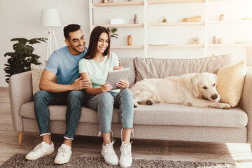 A young couple sits together on a couch, engaged in a conversation while looking at a tablet. A golden retriever relaxes nearby in a bright living room.