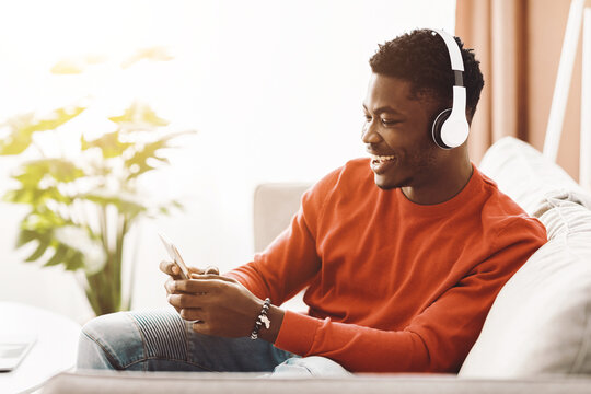 A young man wearing headphones smiles as he listens to music on his smartphone. He is seated on a comfortable couch in a sunny living room, surrounded by indoor plants.