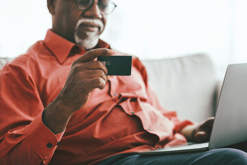 A mature man in a red shirt holds a contactless card in one hand while typing on a laptop. Natural light filters through the window, creating a warm atmosphere in the cozy space.