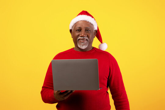 Happy senior African American man in a red sweater and Christmas hat smiles while using his laptop to shop online for holiday deals, set against a vibrant orange background.