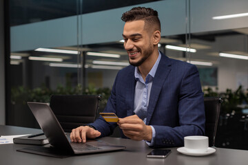 A cheerful middle eastern entrepreneur is sitting at a work desk, typing on a laptop while holding a credit card.