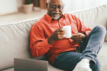 A senior man in a red shirt sits comfortably on a sofa, sipping coffee from a white mug. He appears relaxed and content, enjoying his morning routine. A laptop is nearby.