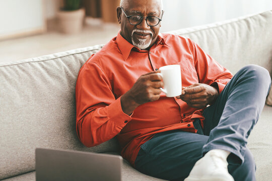 A senior man in a red shirt sits comfortably on a sofa, sipping coffee from a white mug. He appears relaxed and content, enjoying his morning routine. A laptop is nearby.