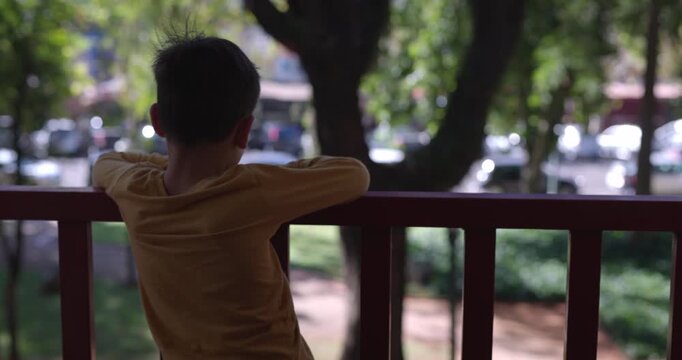 Boy on low city balcony looks out to urban city below - from behind