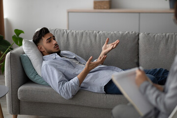 In a psychologist's office, a young Arab man lies on a couch, expressing his feelings. He is...