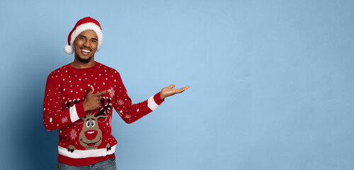 A happy man stands in a red Christmas sweater featuring a reindeer. He wears a Santa hat and points to the side with a cheerful expression, set against a light blue background.