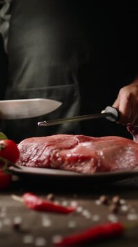 Vertical slow motion video chef sharpening knife behind raw beef meat on table spices and chili pepper in foreground cinematic light dark kitchen atmosphere concept culinary art professional mood