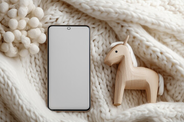 A close-up of a mockup of a smartphone with a white blank screen and a wooden horse figurine lying on a knitted blanket
