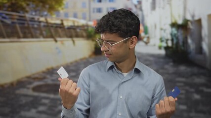 Man in blue shirt holds two creditcard aloft while comparing their chips under sunlight on street lined with urban buildings; hesitation.