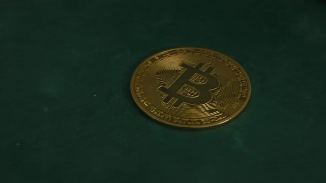 A bitcoin coin lies on a dark green surface as the camera glides past, eventually revealing its mirrored reflection&mdash;suggesting duality, value perception, and the intangible nature of digital currency.