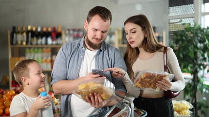 Positive young man and woman with preteen son making purchases in local grocery store, using smartphone to scan barcodes and pay online for packaged cut chicken. Modern shopping concept - Powered by Adobe