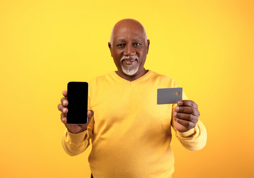 A senior black man holds a credit card in one hand and a cellphone in the other. He stands against a bright orange background, representing modern banking and online shopping concepts.