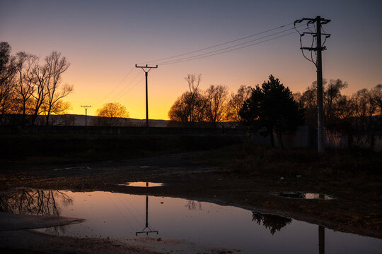 Electric poles and power lines silhouetted at sunset with reflection in a puddle. Rural landscape with warm evening light and calm atmosphere.