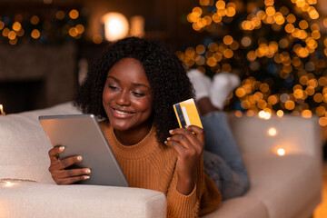 A smiling young African American woman relaxes on her sofa while watching a video on her tablet. She holds a credit card, enjoying online shopping surrounded by a beautifully lit Christmas tree.
